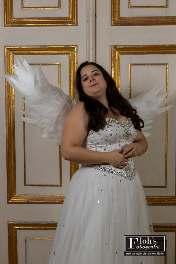 Vici als himmlischer Engel in Burg Hof, elegant vor goldverzierten Türen.