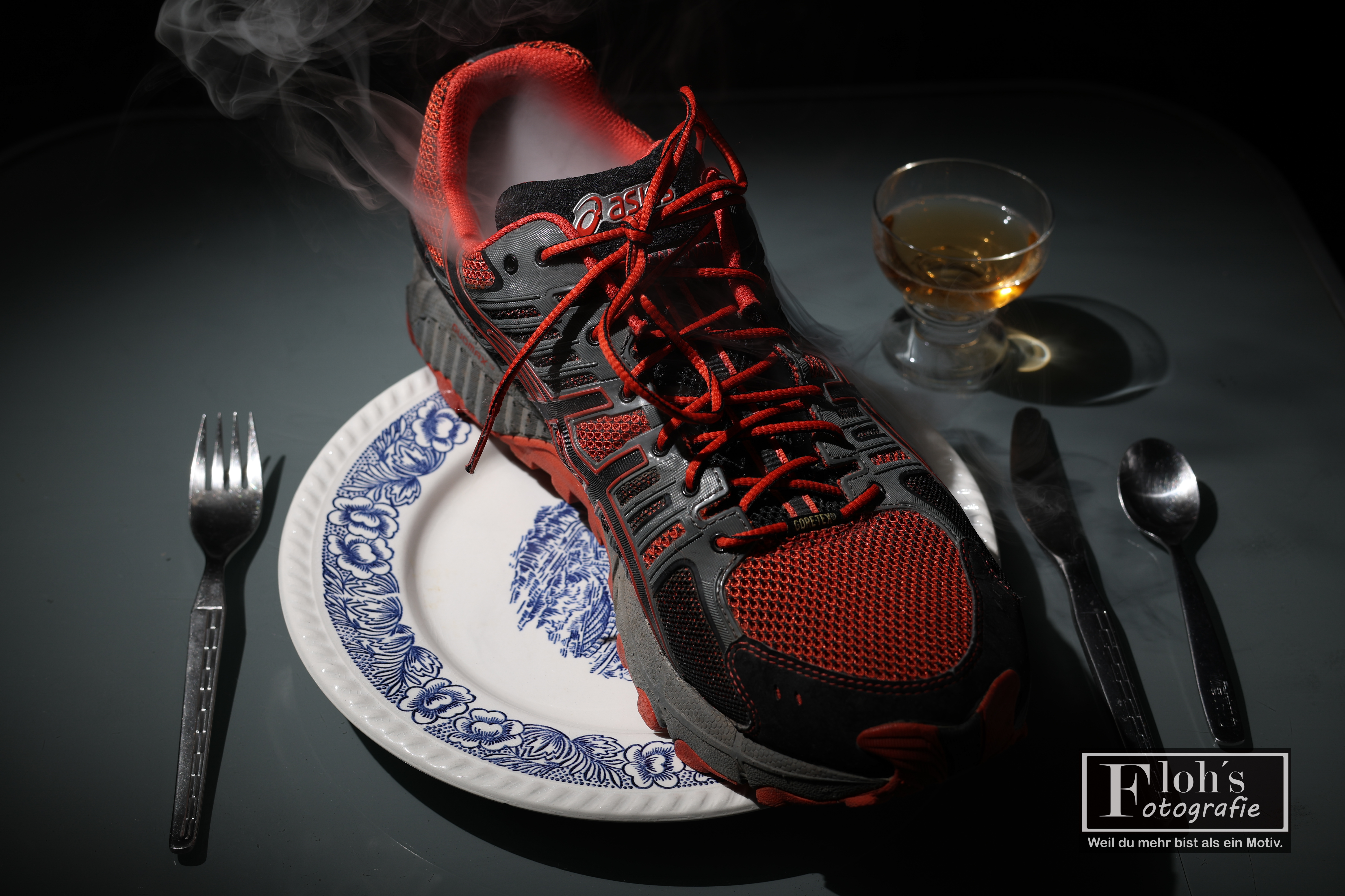 Rot-schwarzer Sportschuh mit aufsteigendem Nebel auf einem weiß-blauen Teller, daneben Besteck und ein gefülltes Glas.