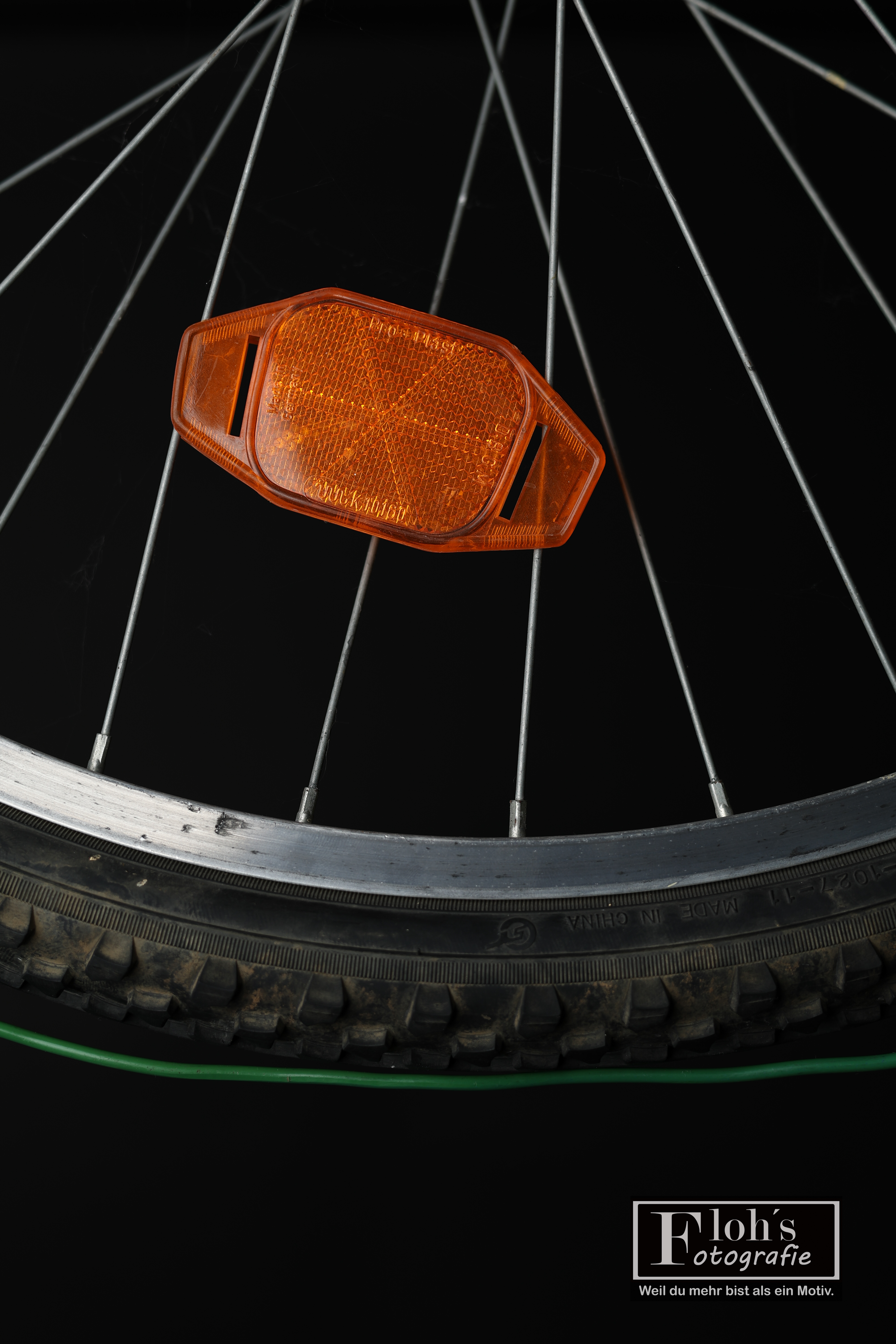 Ausschnitt eines Fahrradrads mit orangefarbenem Speichenreflektor und einem grünen Draht unter dem Reifen vor schwarzem Hintergrund.