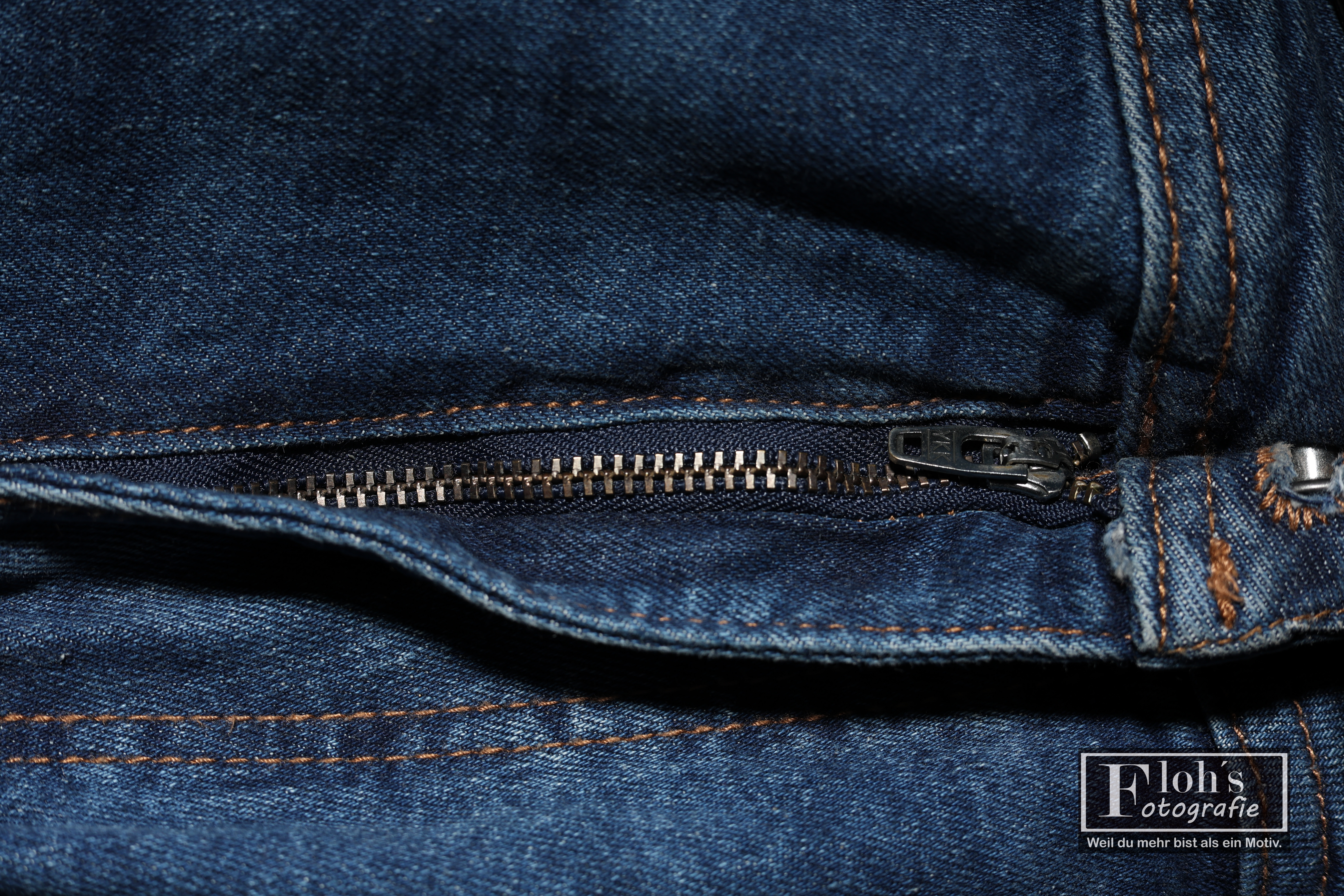 Nahaufnahme eines geschlossenen Metallreißverschlusses in blauem Jeansstoff mit orangebraunen Nähten.