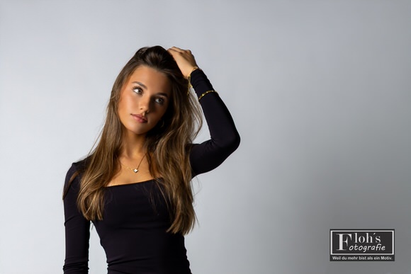 Christina im schwarzen Langarmtop vor hellem Studiohintergrund, mit einer Hand im Haar und seitlich gerichtetem Blick.