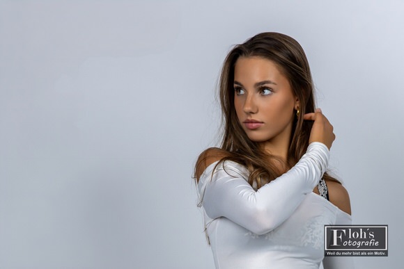 Christina im weißen Oberteil vor hellem Hintergrund, mit einer Hand im Haar und seitlichem Blick aus dem Bild.
