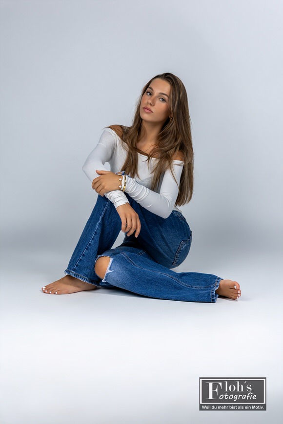 Christina sitzt barfuß in Jeans und weißem Oberteil auf dem Studioboden, mit einem aufgestellten Knie und darauf abgelegten Armen.