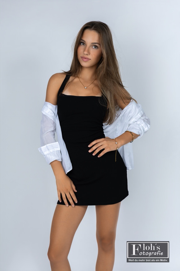 Christina im schwarzen Kleid mit offenem weißem Hemd, einer Hand an der Hüfte und einem leichten Lächeln.