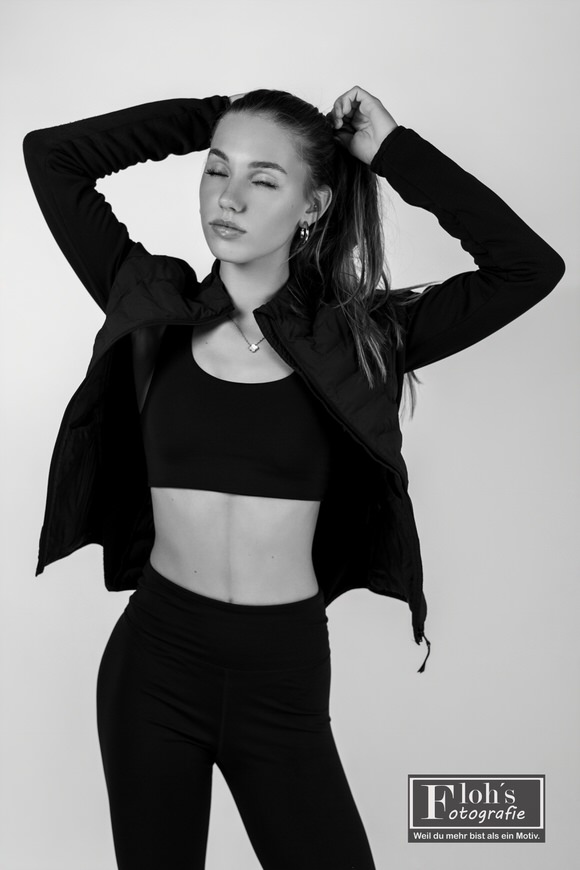 Schwarz-Weiß-Aufnahme von Christina im Sportoutfit mit offener Jacke, geschlossenen Augen und beiden Händen am Pferdeschwanz.