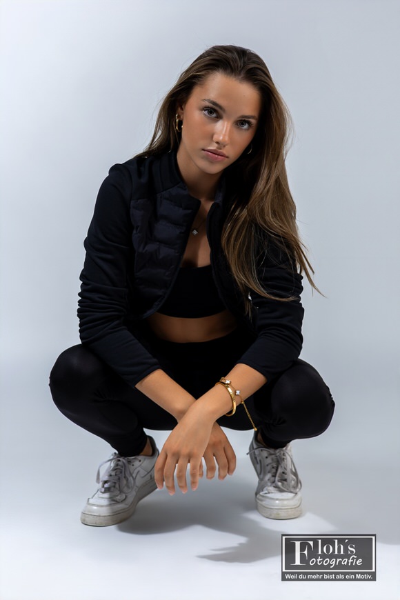 Christina hockt in schwarzem Sportoutfit und weißen Sneakers vor hellem Hintergrund, mit den Unterarmen auf den Knien und Blick in die Kamera.