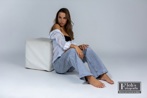 Christina sitzt in Jeans und offenem weißem Hemd auf dem Boden, an einen weißen Sitzwürfel gelehnt und mit angewinkelten Beinen.