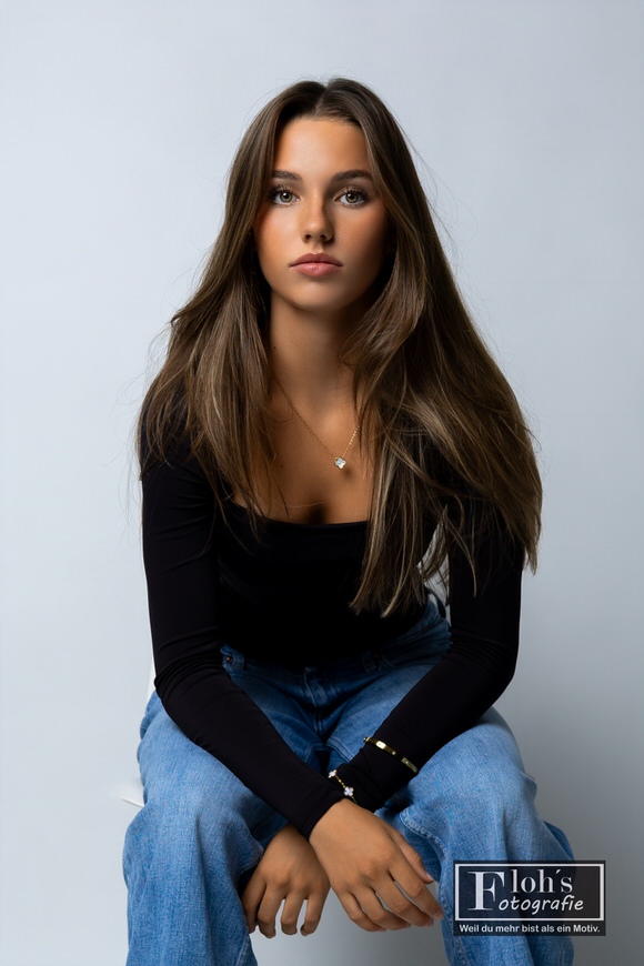 Christina sitzt in Jeans und schwarzem Oberteil auf einem weißen Stuhl, leicht nach vorne gelehnt und mit den Händen zwischen den Knien.