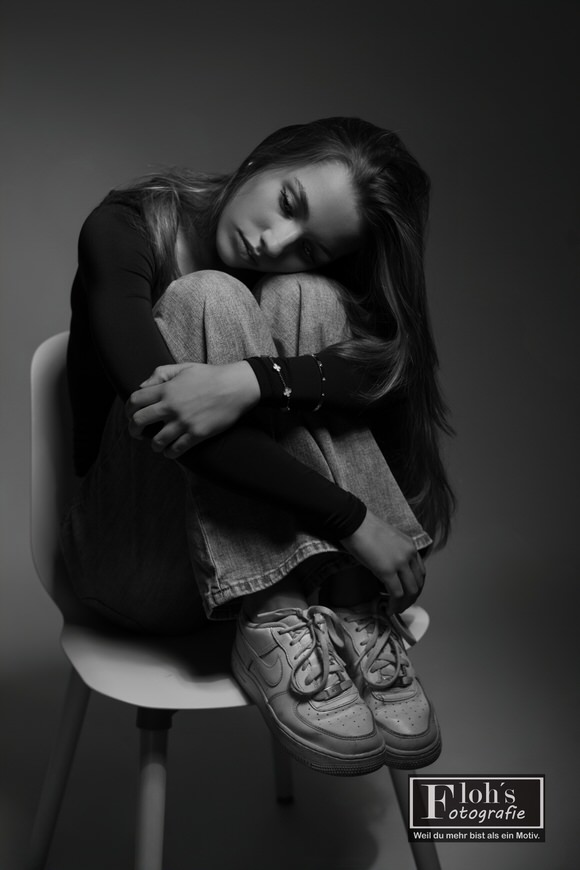 Schwarz-Weiß-Porträt von Christina auf einem Stuhl, mit umschlungenen Knien und seitlich darauf abgelegtem Kopf.