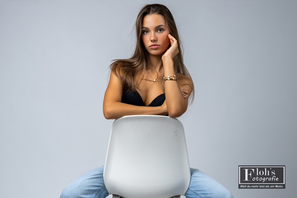 Christina hinter der Lehne eines weißen Stuhls, mit einer Hand an der Wange und direktem Blick in die Kamera.