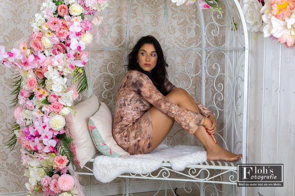 Nicole im roséfarbenen Blumenkleid sitzt mit angezogenen Beinen auf einer weißen, blumengeschmückten Bank und blickt zur Seite.