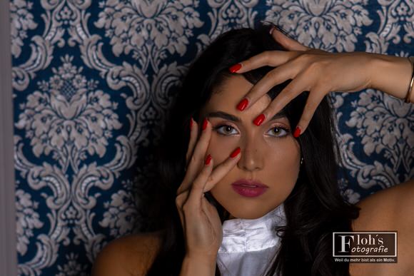 Beauty-Porträt von Nicole vor blau-weiß gemustertem Hintergrund. Ihre Hände mit roten Fingernägeln rahmen Stirn und Wange ein.