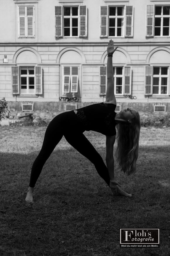 Schwarz-Weiß-Aufnahme von Selina in einer seitlichen Yogapose auf einer Wiese, ein Arm nach oben gestreckt, vor einer Fassade mit Fensterläden.