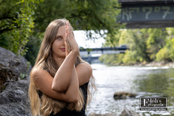 Selina mit verschränkten Armen und aufgerichteten Handflächen vor dem Gesicht, mit der Mur, einer Brücke und grünen Uferbäumen im Hintergrund.
