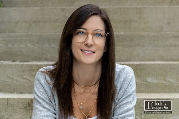 Nahes Portrait von Lisa mit Brille und grauer Strickjacke vor hellen Treppenstufen, mit einem leichten Lächeln zur Kamera.