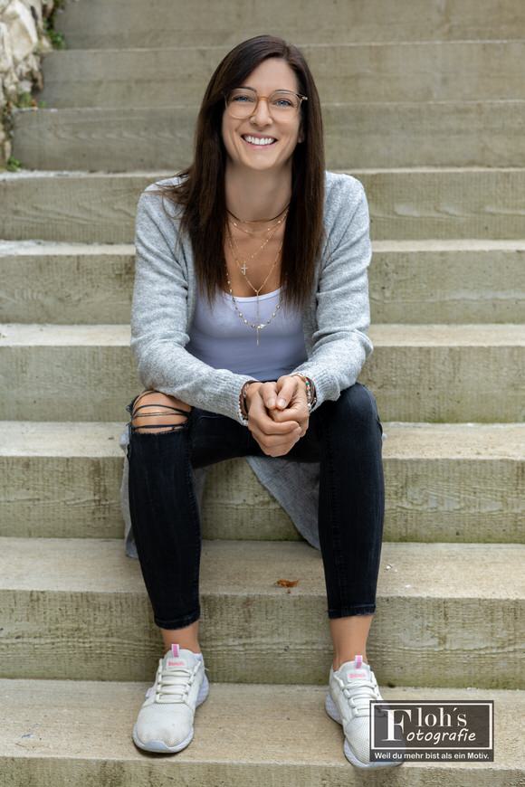 Lisa sitzt lachend auf einer Treppe, trägt eine graue Strickjacke, dunkle Jeans und helle Sneaker und hält die Hände zusammen.