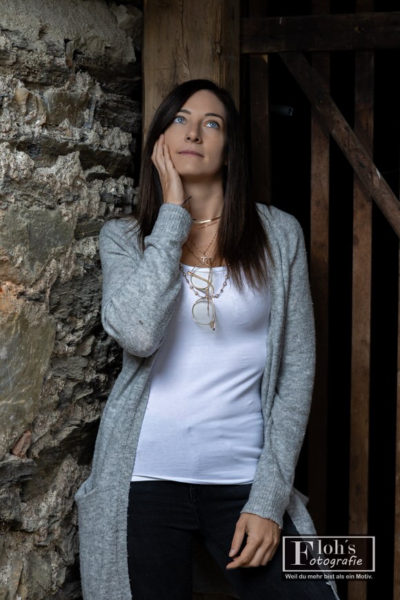 Lisa steht zwischen einer Steinmauer und einem Holztor, legt eine Hand an ihre Wange und blickt nach oben.