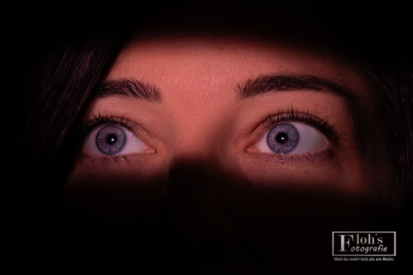 Enge Nahaufnahme von Lisas nach oben gerichteten Augen, beleuchtet von einem rötlichen Lichtstreifen und umgeben von tiefen Schatten.