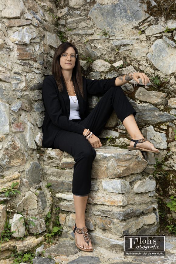Lisa im schwarzen Blazer auf einem steinernen Mauervorsprung, mit aufgestelltem Bein und einem Arm auf dem Knie.