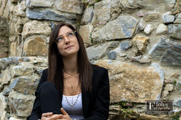 Lisa sitzt mit angezogenem Knie vor einer rauen Steinmauer und blickt durch ihre Brille schräg nach oben.
