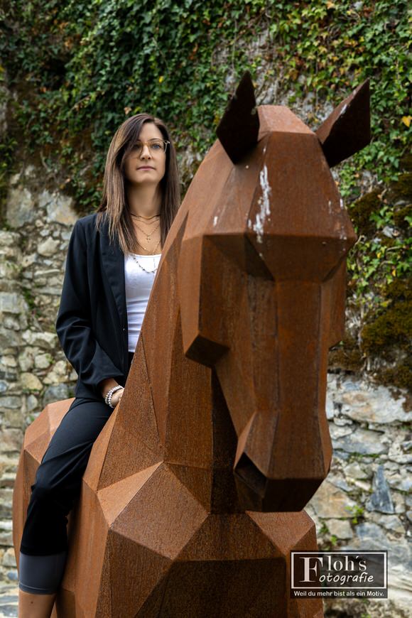 Lisa sitzt auf einer rostfarbenen Pferdeskulptur aus Metall vor einer begrünten Steinmauer der Burgruine Waxenegg.
