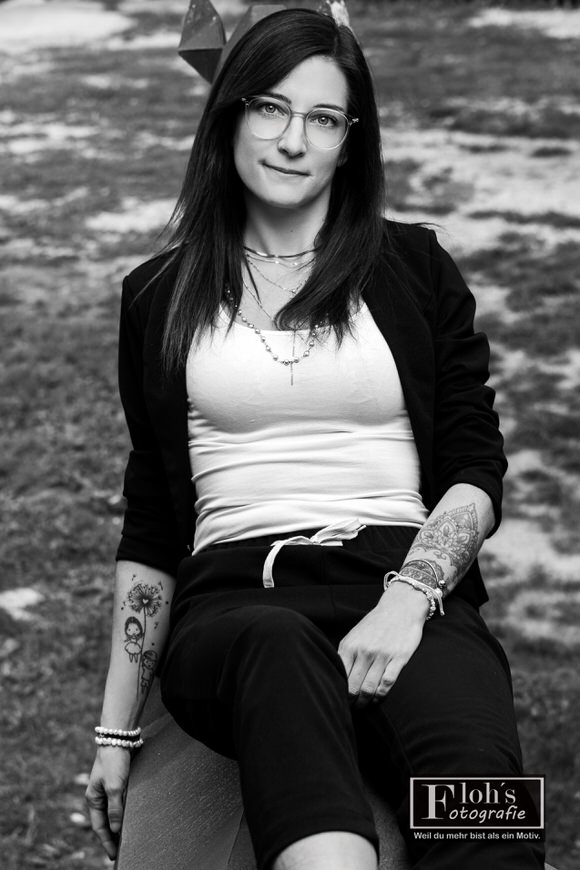 Schwarz-Weiß-Portrait von Lisa im schwarzen Blazer und weißen Top, mit sichtbaren Unterarmtattoos und direktem Blick in die Kamera.