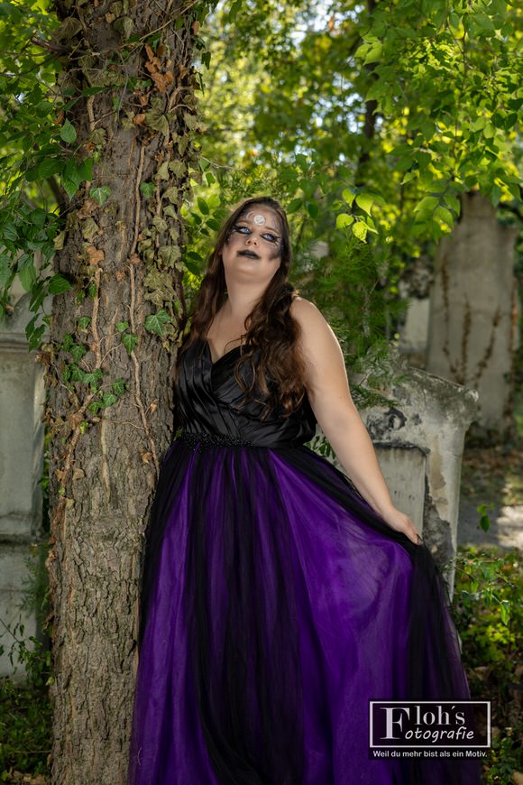 Vici im schwarz-violetten Hexenkleid neben einem efeubewachsenen Baum im St. Marxer Friedhofspark, mit nach oben gerichtetem Blick.