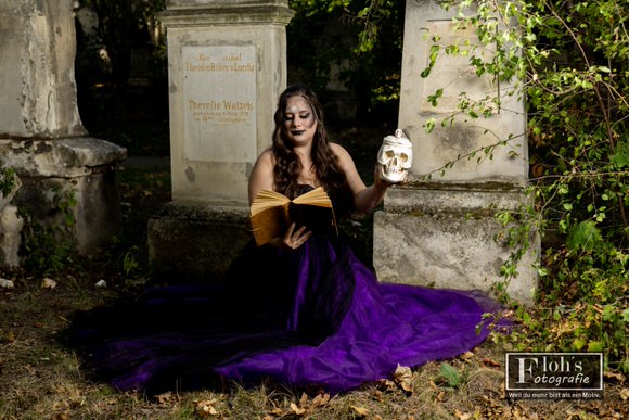 Weite Aufnahme von Vici mit Buch und Totenkopf-Requisite, ihr schwarz-violetter Rock liegt ausgebreitet zwischen den Grabsteinen.