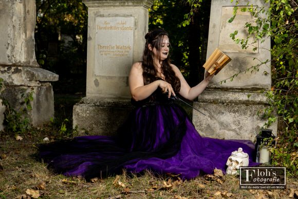 Vici sitzt mit einem Zauberstab vor alten Grabsteinen. Über ihrer geöffneten Hand ist ein Buch zu sehen, daneben stehen Laterne und Totenkopf-Requisite.