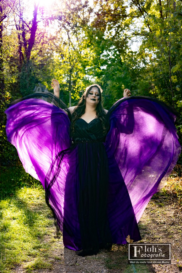 Vici mit erhobenen Armen und weit auffliegendem violettem Kleiderstoff, durch den das Sonnenlicht zwischen den Bäumen scheint.