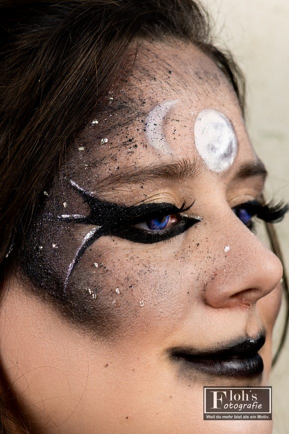 Detailaufnahme von Vicis Hexen-Make-up mit geschwungenen schwarzen Augenlinien, Glitzer und weißen Mondsymbolen auf der Stirn.