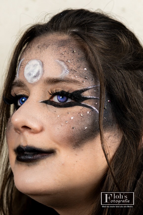 Nahes Dreiviertelportrait von Vici mit blau-violetten Augen, schwarzen Lippen, schimmernden Make-up-Details und Mondsymbolen auf der Stirn.