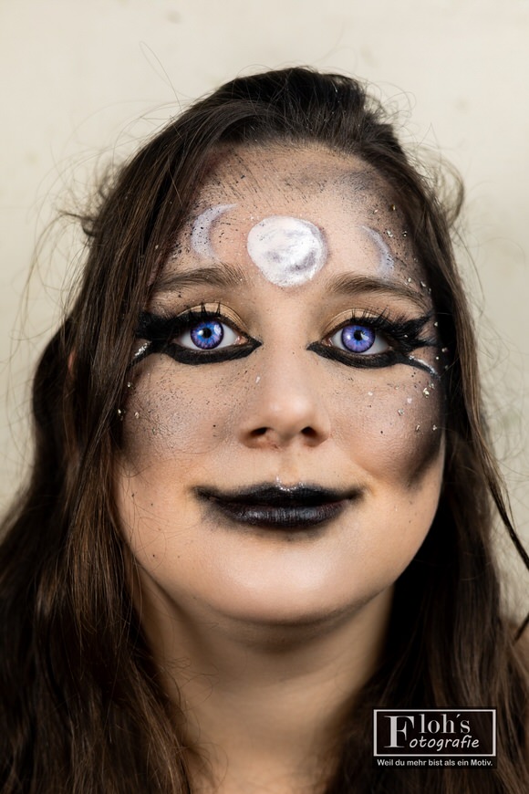 Frontalportrait von Vici mit direktem Blick, schwarzen Lippen, markantem Augen-Make-up und hellen Mondsymbolen auf der Stirn.
