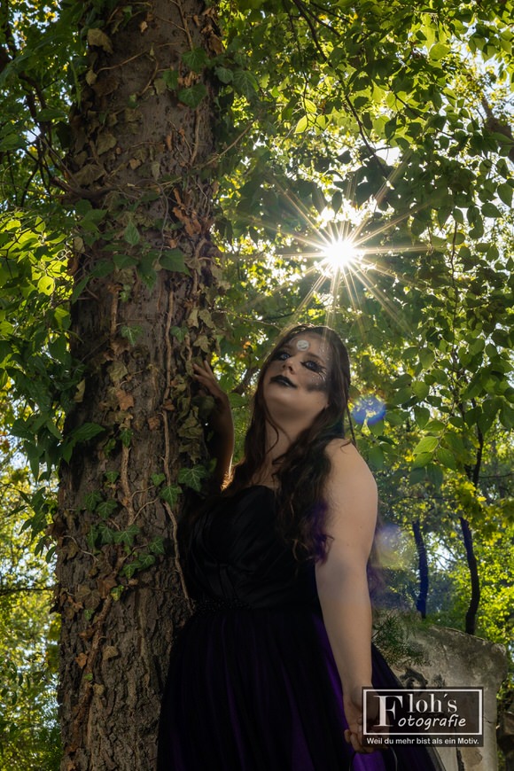 Vici neben einem efeubewachsenen Baum, während die Sonne sternförmig durch das Blätterdach über ihrem Kopf scheint.