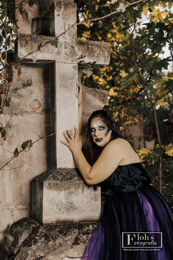 Vici lehnt im schwarz-violetten Hexenkleid an einem großen steinernen Kreuz und blickt direkt in die Kamera.