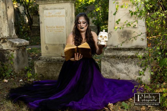 Vici sitzt zwischen alten Grabsteinen und hält ein aufgeschlagenes Buch sowie eine weiße Totenkopf-Requisite.