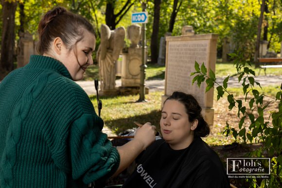 Make-up-Artistin Jessi schminkt Vici auf einer Bank unter Bäumen, während das Model mit geschlossenen Augen einen schwarzen Schminkumhang trägt.