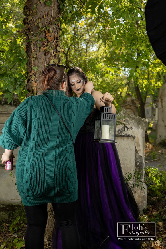 Jessi korrigiert Vicis Styling neben einem Baum, während das Model im schwarz-violetten Kleid eine Laterne hält.