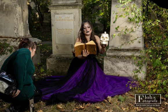 Jessi richtet den ausgebreiteten violetten Rock, während Vici mit einem offenen Buch und einer Totenkopf-Requisite zwischen alten Grabsteinen sitzt.