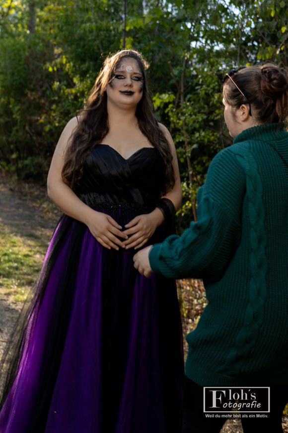 Vici steht mit Hexen-Make-up und schwarz-violettem Kleid zwischen Bäumen, während Jessi ihr im grünen Pullover gegenübersteht.