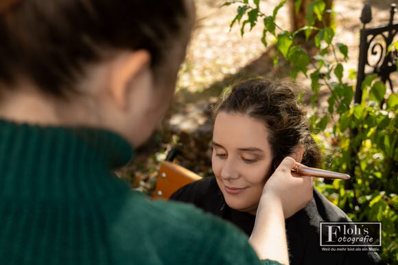 Blick über Jessis Schulter, während sie mit einem Pinsel Make-up auf Vicis Wange aufträgt.