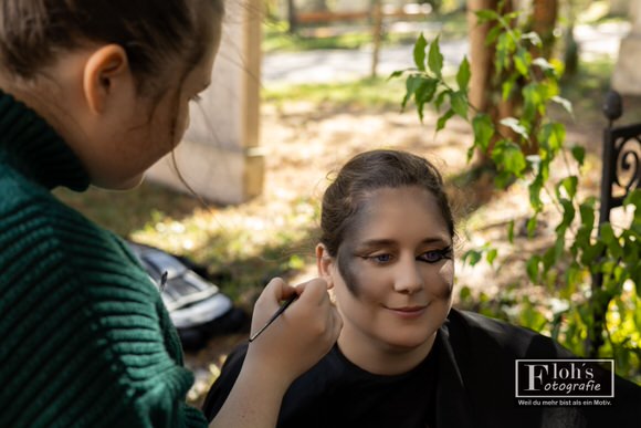 Jessi gestaltet Vicis dunkles Augen-Make-up mit einem feinen Pinsel, während das Model auf der Bank sitzt.