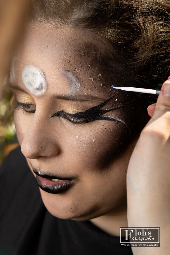 Nahaufnahme von Vicis Hexen-Make-up mit schwarzen Augenlinien, Glitzer und Mondsymbolen, während ein feiner Applikator Details an der Schläfe ergänzt.